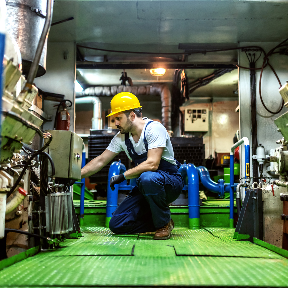 Worthy caucasian mechanic in overalls and with helmet kneeling inside ship and repairing engine.